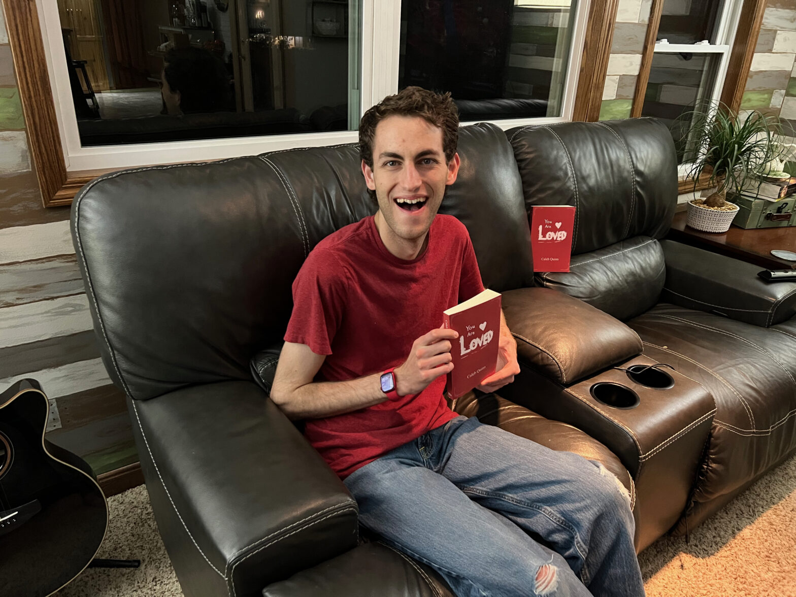Author Caleb Quinn sits on a couch holding his book, "You Are Loved."